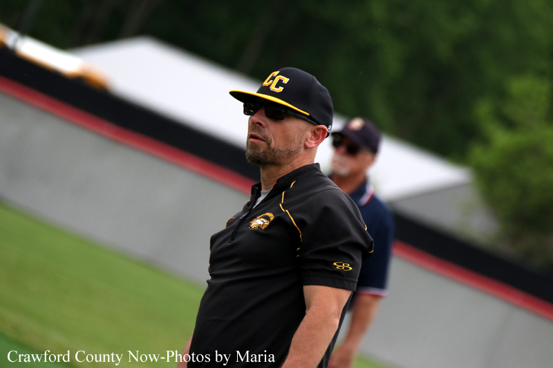 Male coach wearing a black polo and cap with yellow accents, standing on a grassy field and looking off to the side.