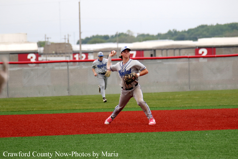 Baseball player in gray uniform prepares to throw on a red infield, with another fielder running in the background against a chain-link fence.