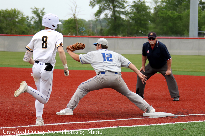 Baseball scene: a runner wearing white with number 8 sprints toward the base as a fielder in gray with number 12 lunges to tag at the base; an umpire watches from behind on the infield red dirt.