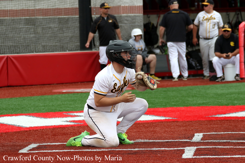 Baseball catcher kneeling on a red infield, wearing a white and gold Eagles uniform and black helmet, glove ready for a throw.