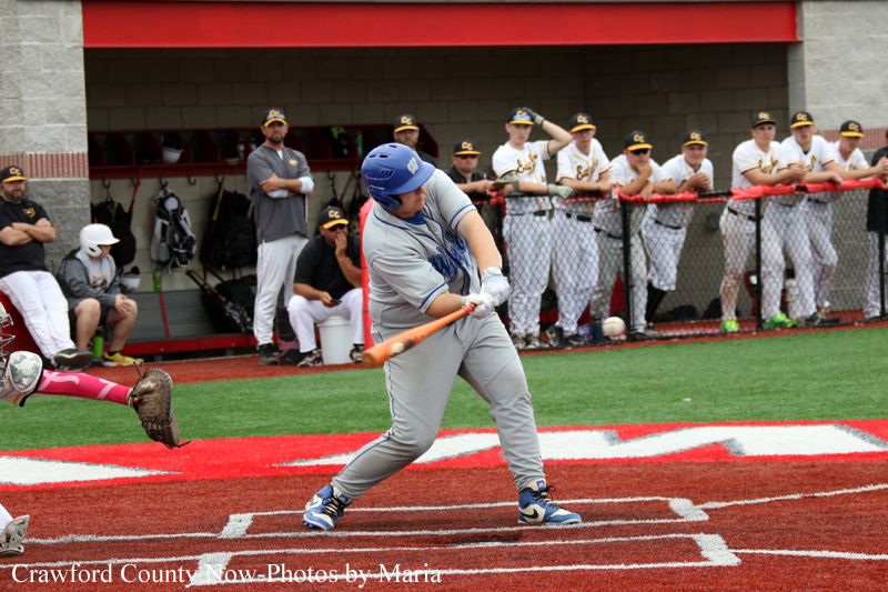 Baseball batter in gray uniform swings a bat on a red infield, with a catcher nearby and teammates watching from the dugout behind the fence.