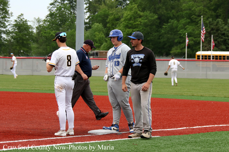 Baseball players near first base on a sunny field, with an umpire walking by and teammates waiting on the dirt and grass.