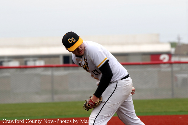 Baseball pitcher in white uniform with black and yellow cap winds up on the mound during a game, glove ready, fence and field behind him.