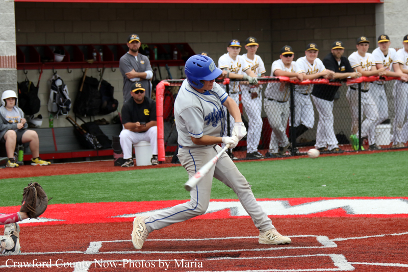 Baseball batter in a blue helmet swings at a pitch at home plate while teammates and coaches watch from the dugout and fence in the background.