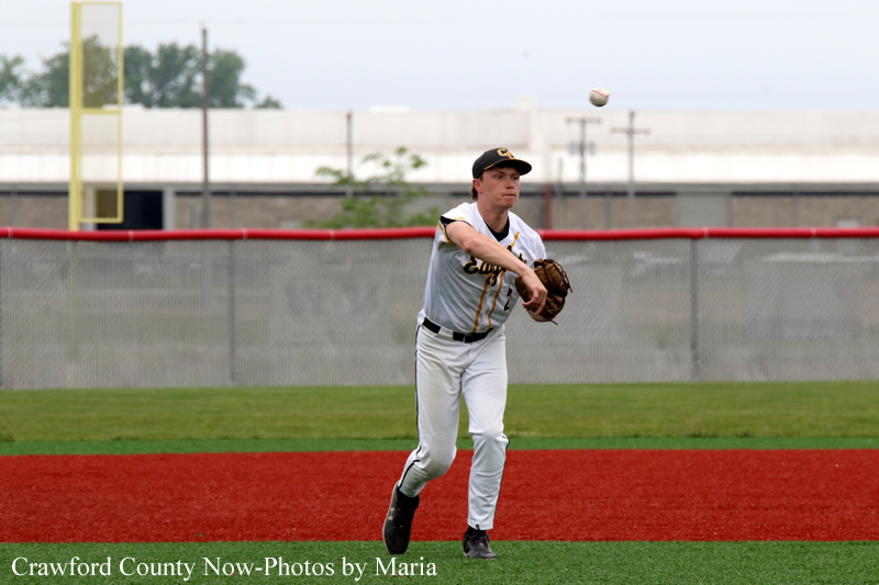 Baseball player in a white uniform throws a ball on the red infield with a chain-link fence in the background.