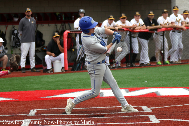 Baseball batter in gray uniform swinging at a pitch at home plate, with teammates watching from the dugout in the background