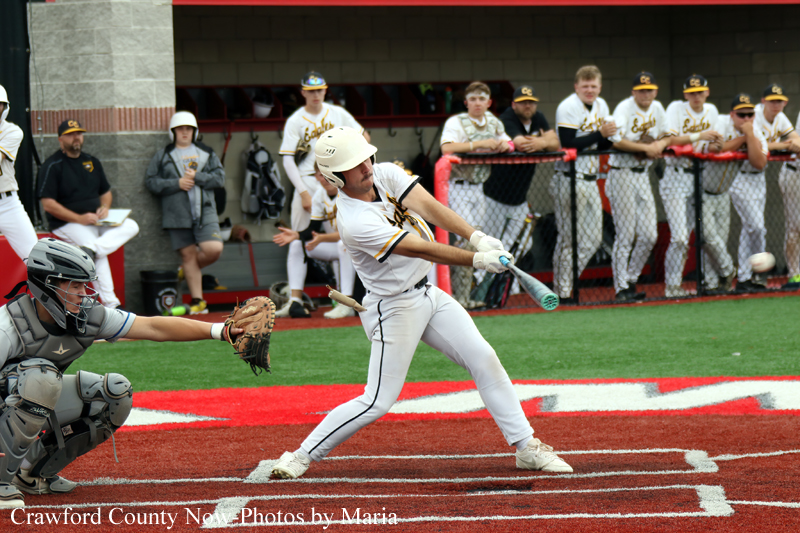 Baseball batter in white with a helmet swings at a pitch as the catcher extends his glove behind him on a red-dirt field with teammates watching from the dugout.