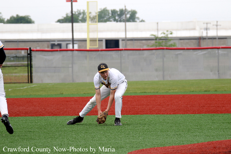 Baseball player in white uniform crouches to field a ground ball on a green and red turf field with a backstop fence behind him to the right.
