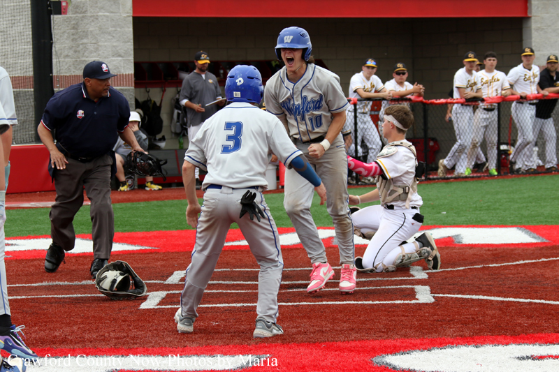 Baseball players celebrate at home plate: a gray-uniform batter jumps in triumph while a teammate in white stands nearby and a catcher kneels, with an umpire and dugout in the background.