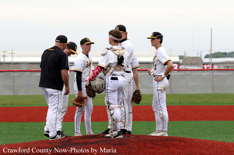 Group of young baseball players in white uniforms huddling on the pitcher's mound with a coach in black nearby; bottom caption reads Crawford County Now-Photos by Maria.