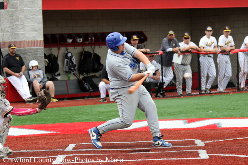 Baseball batter in gray uniform and blue helmet swings at a pitch near home plate, with teammates watching from the dugout in the background.