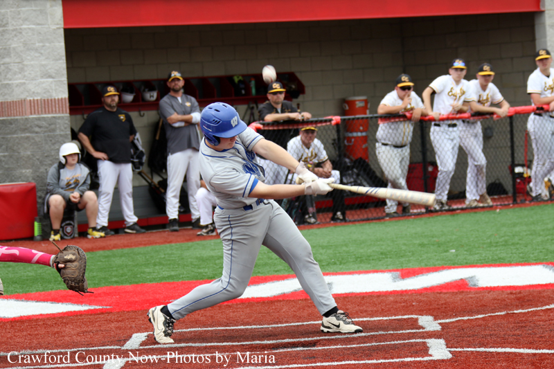 Baseball batter in gray uniform and blue helmet swings at a pitch near home plate, with teammates watching from the dugout in the background.