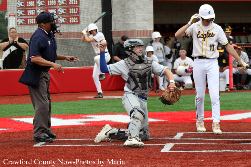 Baseball catcher kneels at home plate with glove raised, signaling a play as the umpire and a batter watch nearby.
