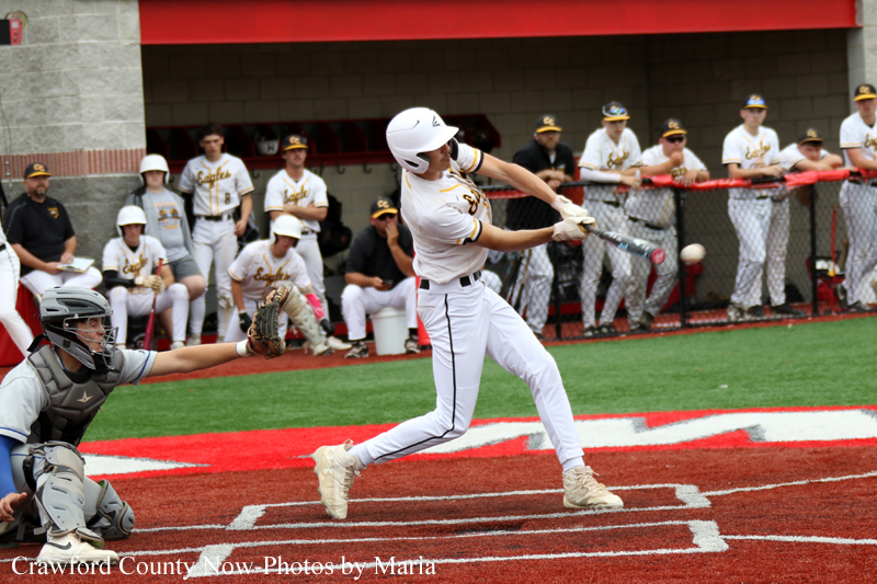 Baseball batter in white uniform swings at a pitch as catcher crouches behind home plate; teammates watch from the dugout.