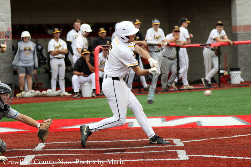 Baseball batter in a white uniform swings at a pitch near home plate; catcher crouches to the left and teammates watch from the dugout in the background.