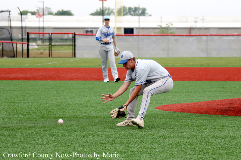 Baseball player in gray uniform dives to field a ground ball, glove low to the turf; another player watches from the infield in the background.