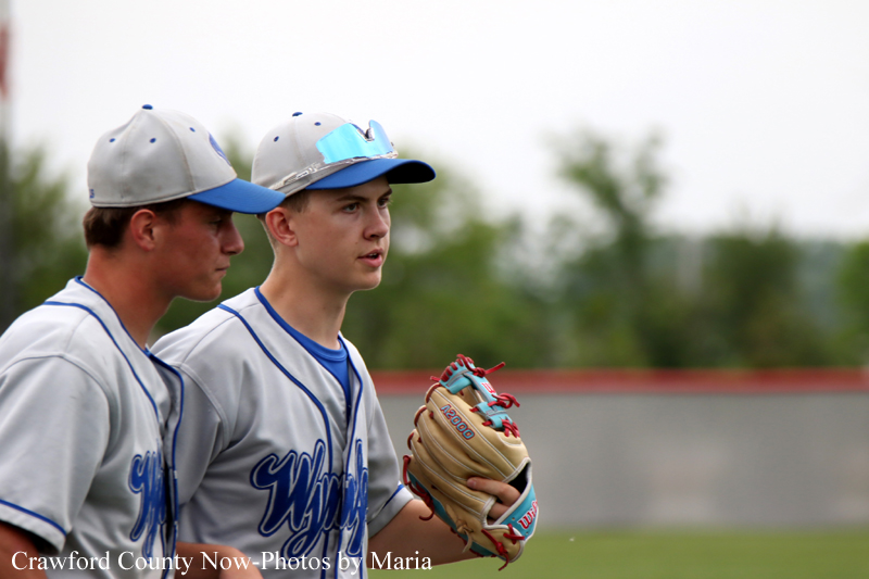 Two youth baseball players in gray jerseys and blue caps stand on a baseball field, the front player holding a glove and looking ahead.