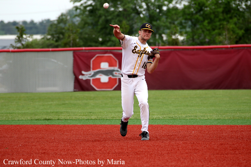 Baseball pitcher in a white uniform mid-throw on a red-dirt infield with a team banner in the background.
