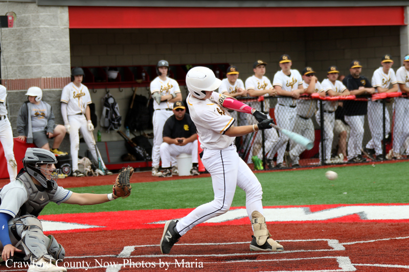 Baseball batter in white uniform swings at a pitch as the catcher crouches behind home plate and teammates watch from the dugout behind the fence.