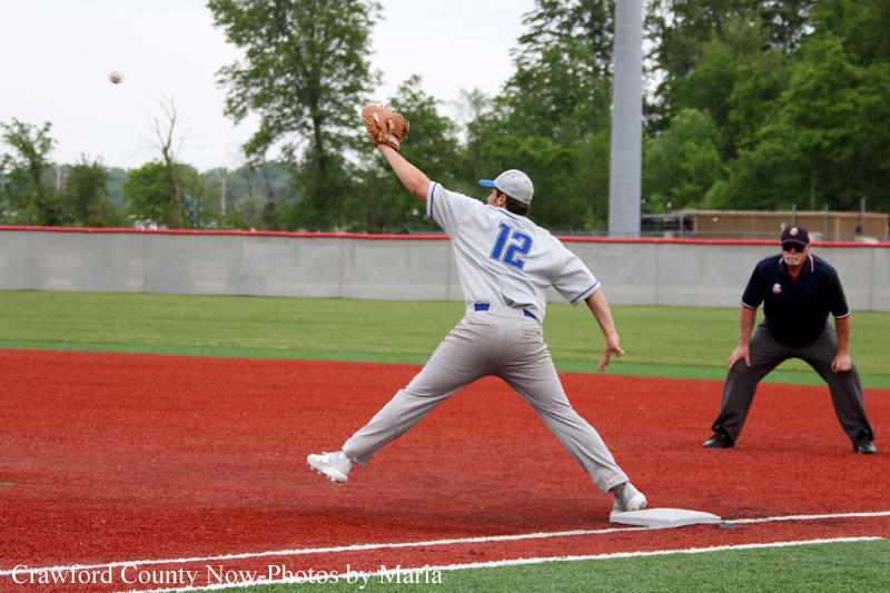 Baseball pitcher wearing number 12 delivers a pitch from the mound on a red infield, with an umpire in the background.