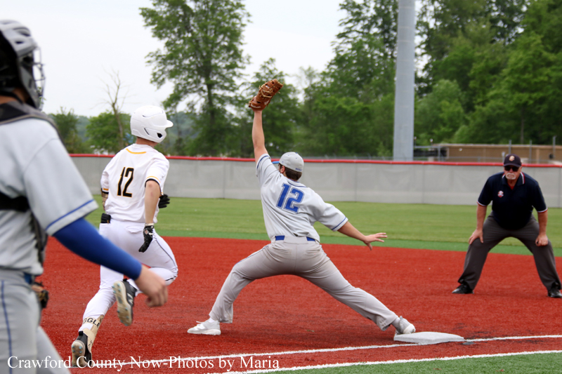 Baseball scene: a gray-uniformed pitcher (no. 12) leaps and throws from the mound as a white-uniformed baserunner advances toward second base; umpire watches nearby on the red infield dirt.