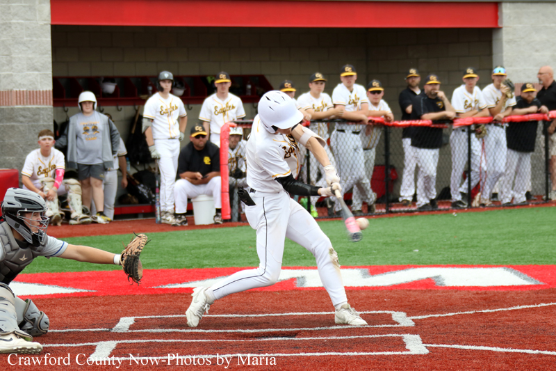 Baseball batter in white swings at a pitch at home plate as catcher reads the ball; dugout and spectators watch from behind a chain-link fence.