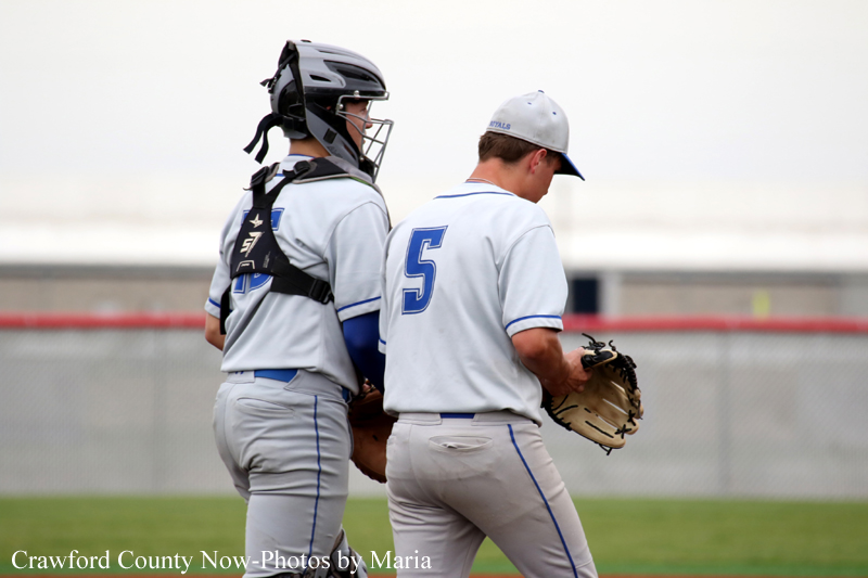 Two baseball players walk side by side on a field; a catcher in a black helmet and chest protector beside a pitcher wearing number 5 on his back in gray uniforms.