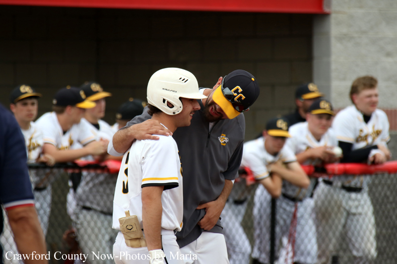 Baseball player in white uniform and helmet receives coaching from a teammate near the dugout fence, others watch in the background.
