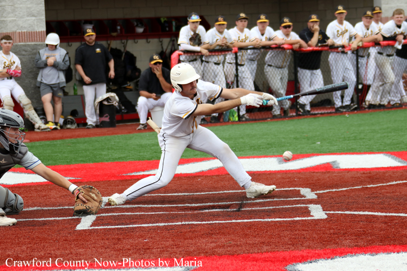 Baseball batter in white uniform swings at a pitch at home plate, with catcher crouched behind and teammates watching from the dugout.