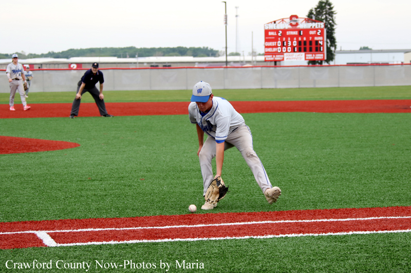 Baseball fielder in white uniform scoops a ground ball near the first-base line on a turf field, with a scoreboard and players in the background.