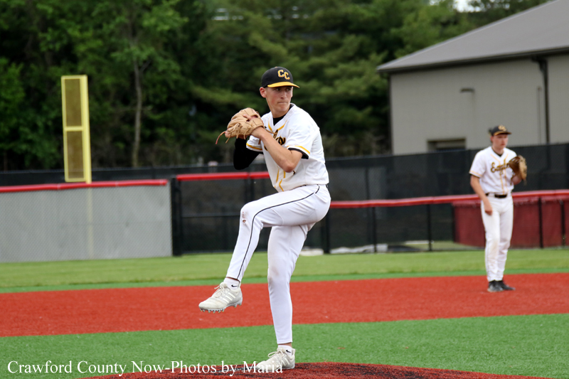 Baseball pitcher in a white uniform mid-pitch on the mound, glove raised, with a teammate in the background on the field.