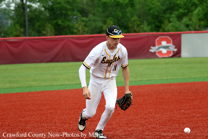 Youth baseball player in white and gold Eagles uniform fields a ground ball on a red dirt infield, glove ready, ball nearby