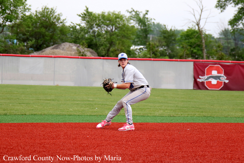 Young baseball pitcher in a gray uniform crouches on the mound, glove up, ready to throw on a red infield.