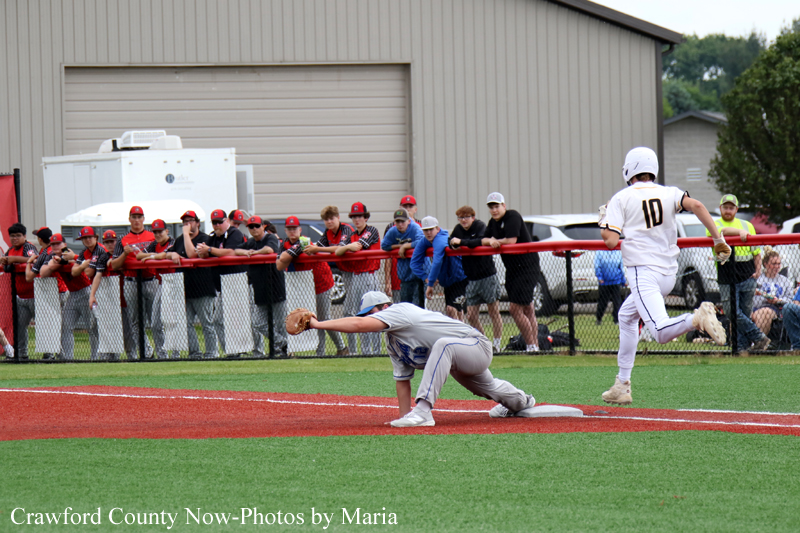 Baseball action at home plate: a fielder reaches for a tag as a runner slides in, with spectators along the chain-link fence behind them.
