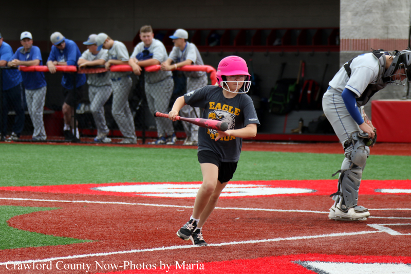 Young girl in a pink helmet runs with a bat during a youth softball game, with a catcher and umpire nearby on the field.