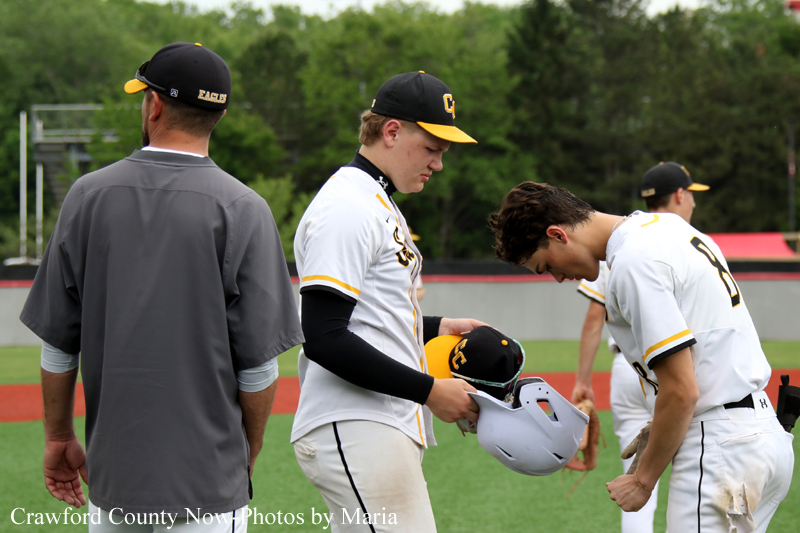 Baseball players in white uniforms with black and yellow caps stand on the field; one player holds a helmet as another leans forward, while a coach in a gray jacket faces away.