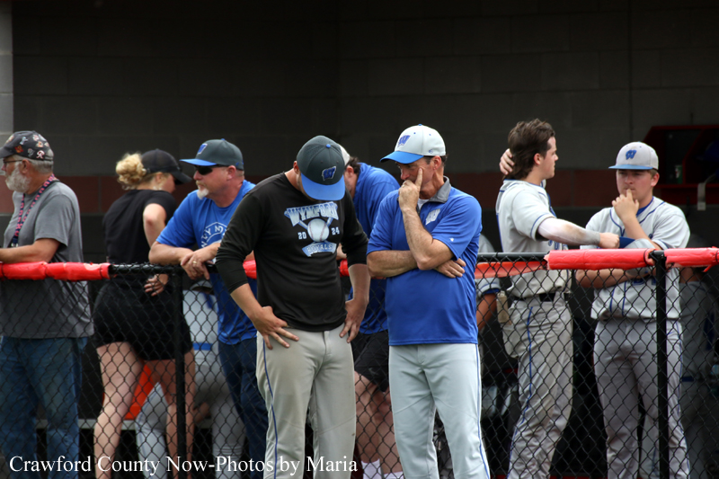 Group of men and a woman leaning on a chain-link fence, watching a baseball game from the dugout area.