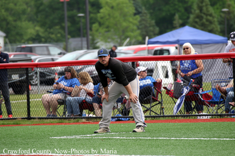 Baseball player in a black shirt and gray pants crouches in the infield, ready to field, with spectators seated behind a chain-link fence.