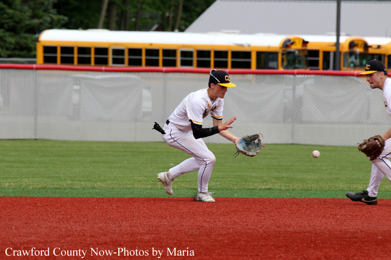 Baseball infielder in a white uniform lunges with his glove to field a ground ball; another fielder watches nearby, on a grassy field with a red infield.