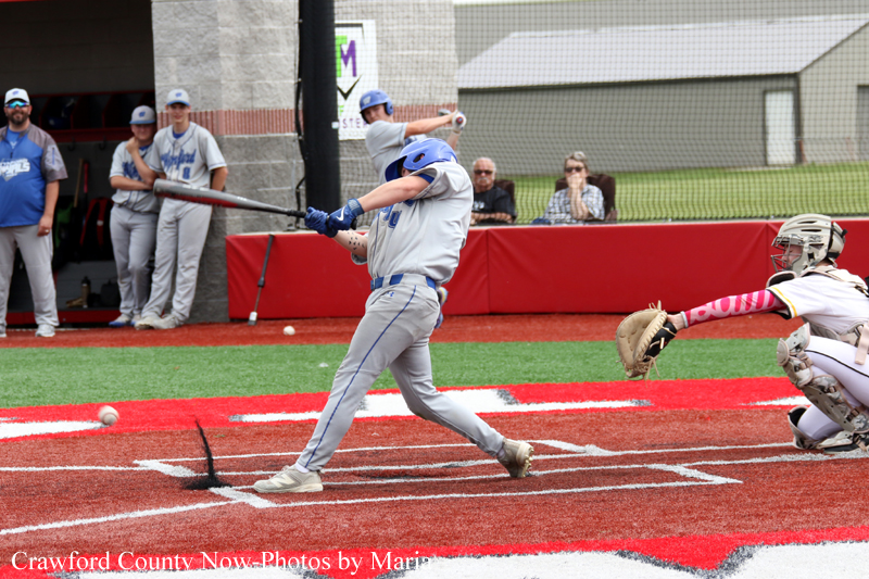 Baseball batter swings at a pitch near home plate as the catcher dives to block the ball at a red-fenced field edge.