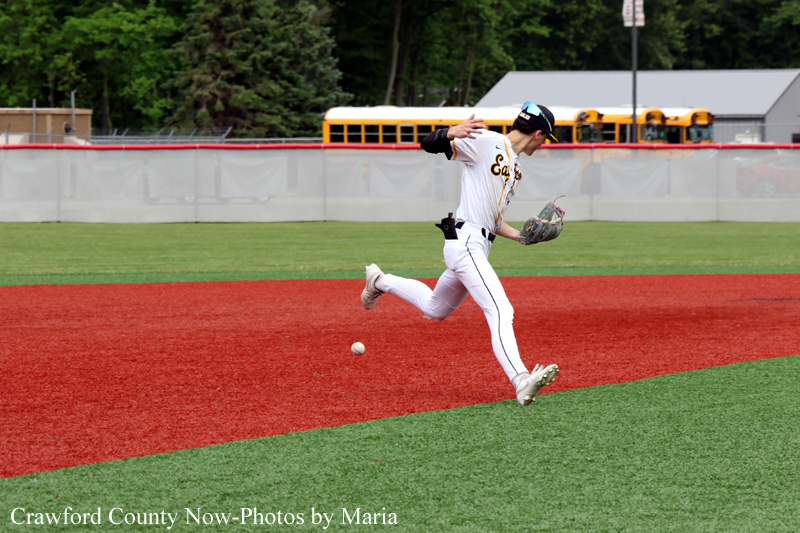 Baseball pitcher in a white uniform throws a pitch on a red infield, with a ball nearby and a bus visible in the outfield background.