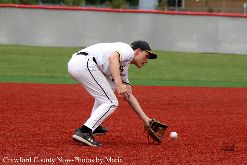 Baseball fielder in a white uniform crouches to field a ground ball on a red infield dirt.