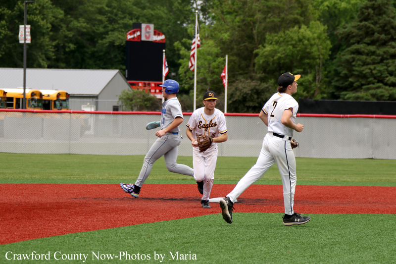 Three youth baseball players sprint across a red-dirt infield with green grass, one wearing a white 'Eagles' uniform, others in gray and white, during a game near the outfield fence and trees.
