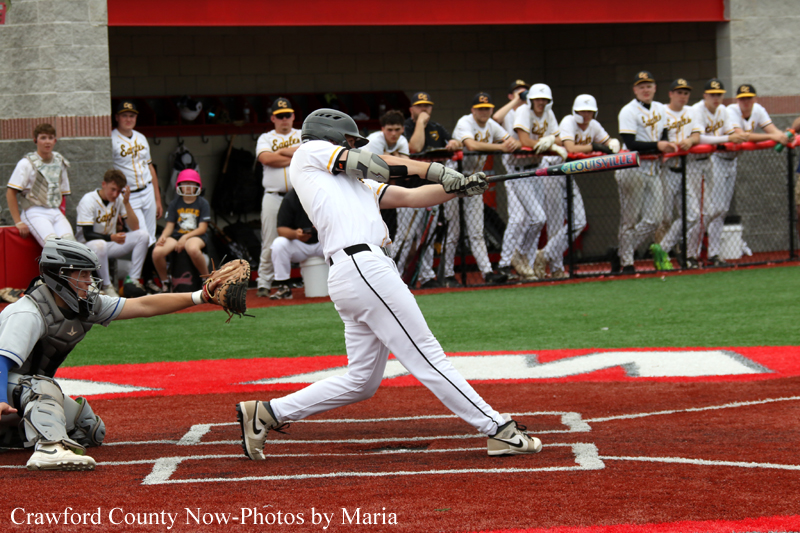 Baseball batter in white swings at a pitch as the catcher crouches behind home plate; crowd and dugout visible in the background.
