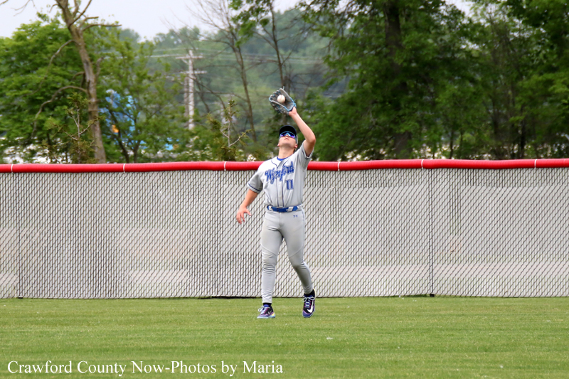 Baseball player in a gray uniform and blue cap reaches up to catch a fly ball near the outfield fence.
