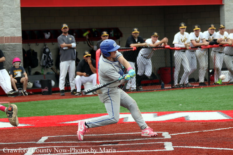 Baseball batter in a gray uniform and blue helmet swings at a pitch on a red dirt infield, with teammates watching from the dugout and behind a chain-link fence.