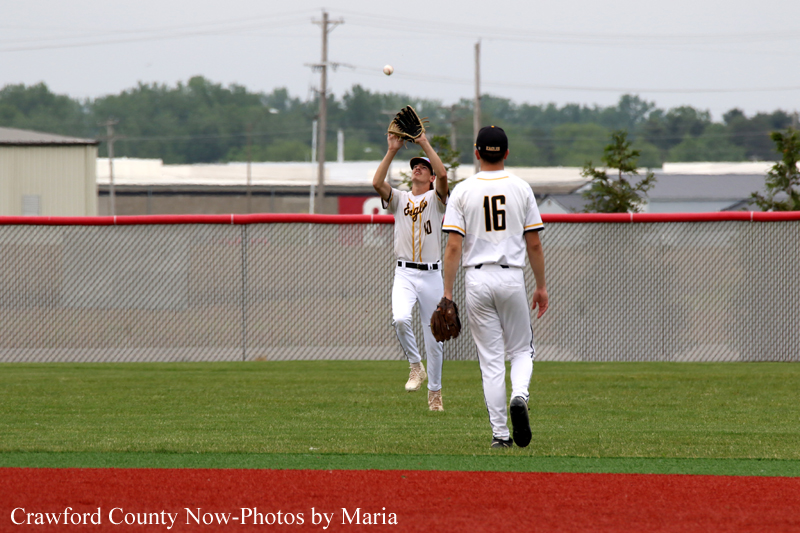 Two baseball players in white uniforms on a grass field; one leaps with glove up to catch a ball, while a teammate stands nearby.