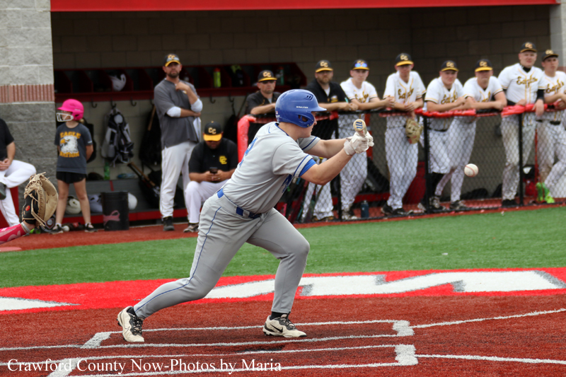 Batter in a gray uniform and blue helmet swings at a pitch at home plate as teammates watch from the dugout behind him on a baseball field.