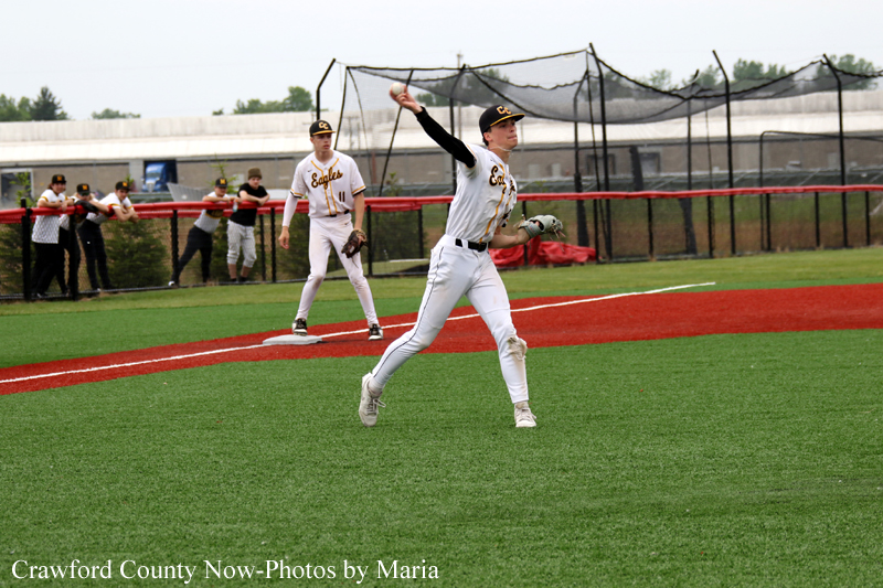 Baseball game action: pitcher in white uniform winds up to throw on a green field with teammates and dugout in the background
