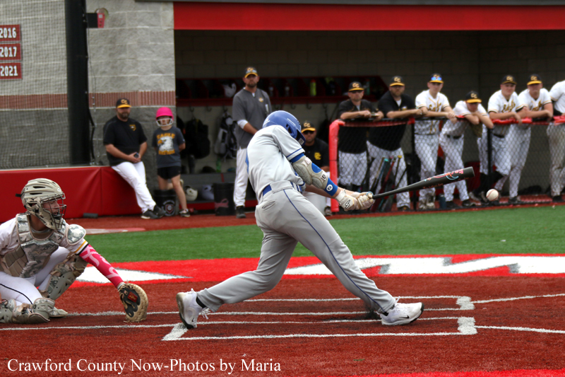 Baseball batter in gray uniform swings at a pitch at home plate; catcher in white gear crouches behind him, while teammates watch from the dugout in the background.